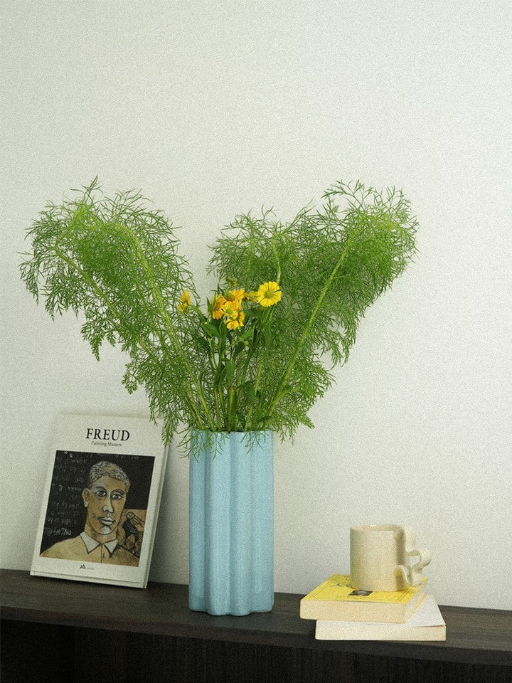 Cosik Home light blue ceramic fluted flower vase with wavy silhouette and accordion texture, holding green foliage and yellow flowers.